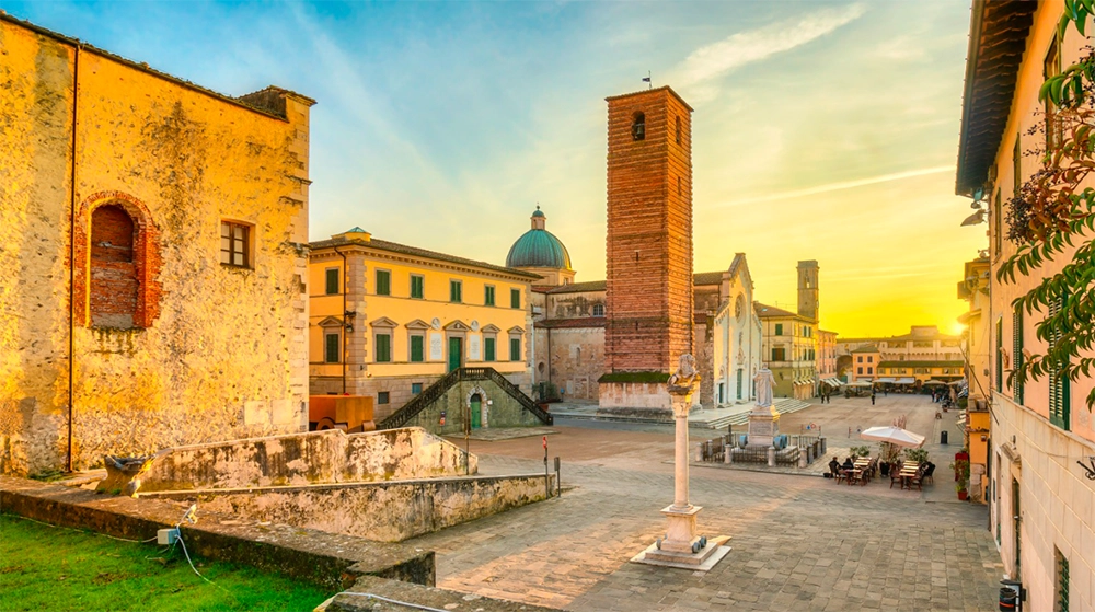  Vista panoramica della Piazza del Duomo di Pietrasanta al tramonto, con il campanile di San Girolamo e la chiesa di San Martino sullo sfondo. Le antiche facciate dei palazzi storici si affacciano sulla piazza, mentre i tavolini all'aperto creano un'atmosfera accogliente e rilassata.