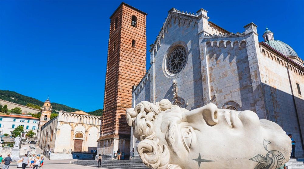 Vista di Piazza del Duomo a Pietrasanta con il campanile di San Girolamo e la facciata della Chiesa di San Martino. In primo piano, la grande scultura del volto di un uomo, opera di arte contemporanea, che si staglia contro il cielo azzurro.