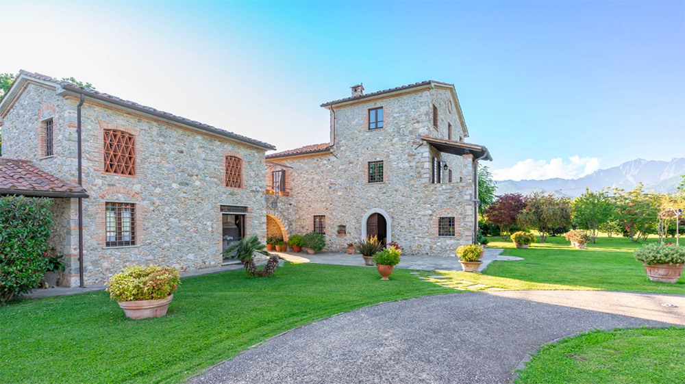 Vista esterna di una villa a Pietrasanta, una residenza in pietra con dettagli in mattoni, circondata da un ampio giardino verde. Sullo sfondo, si stagliano le montagne che creano una cornice naturale per questa affascinante villa toscana.