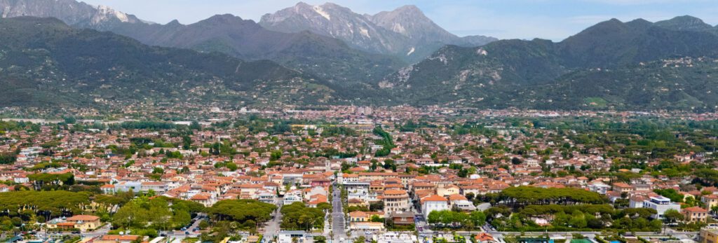 Vista aerea di Forte dei Marmi con la spiaggia attrezzata, il pontile e il centro cittadino, incorniciati dalle Alpi Apuane, simbolo della Versilia più esclusiva.
